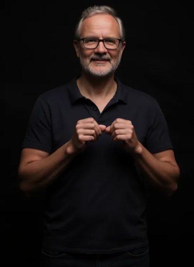 Ekkehard Schmider, wearing glasses and a dark shirt, standing with hands clasped, smiling against a black background during the free ai course.